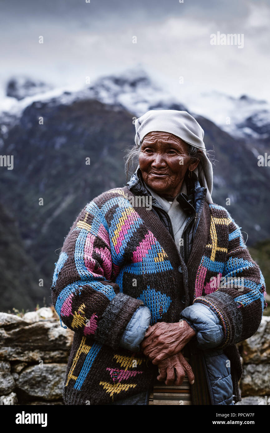 An elderly Sherpa woman standing in a colorful coat before the mountains on a ridge line near Namche Bazaar, Solu Khumbu, Nepal Stock Photo