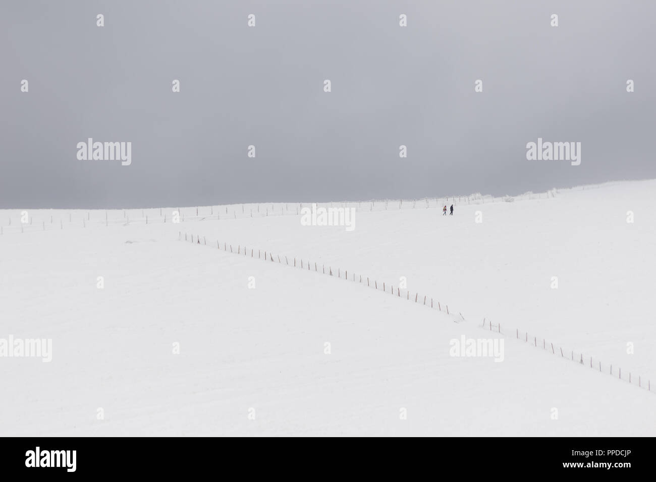 A very minimalistic view of two distant people over a mountain covered by snow, near a fence Stock Photo