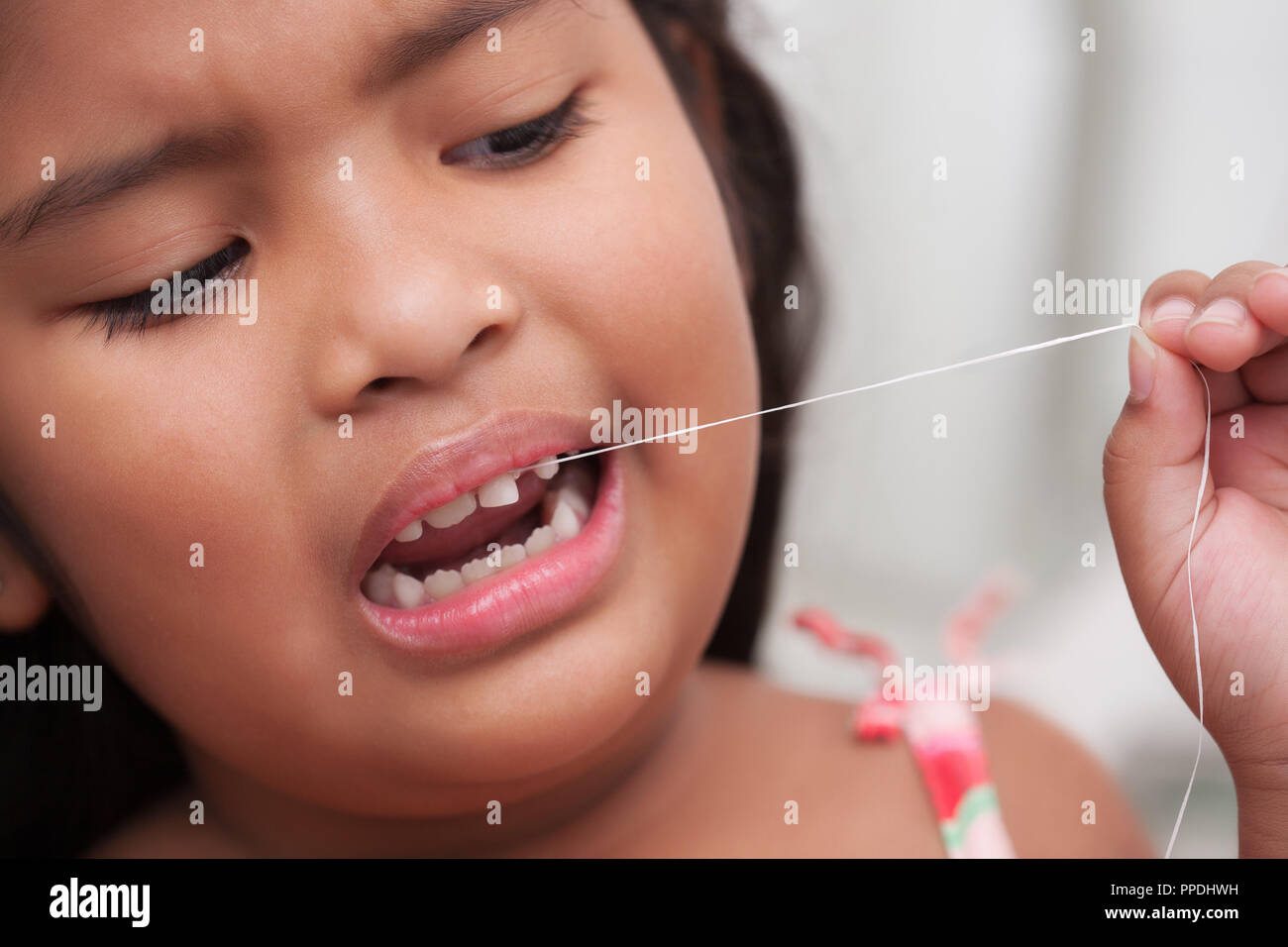 Scared little girl about to pull out her own front milk tooth, using floss string tied to the loose tooth Stock Photo