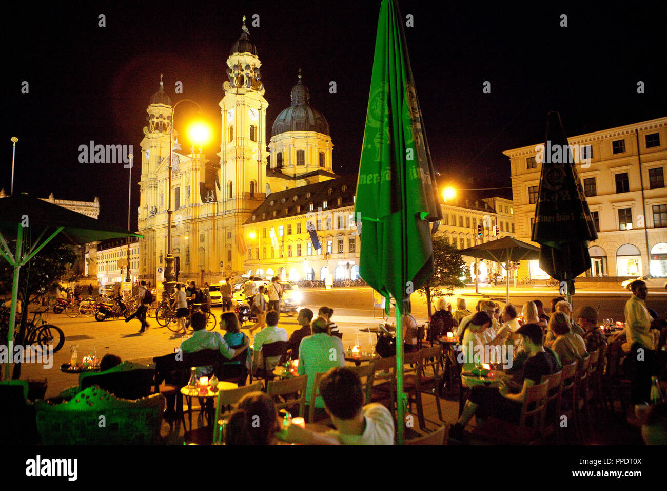 Guests sit on the terrace of Cafe Tambosi on Odeonsplatz on a warm summer evening. In the background, the illuminated Theatinerkirche (Theatine Church). Stock Photo