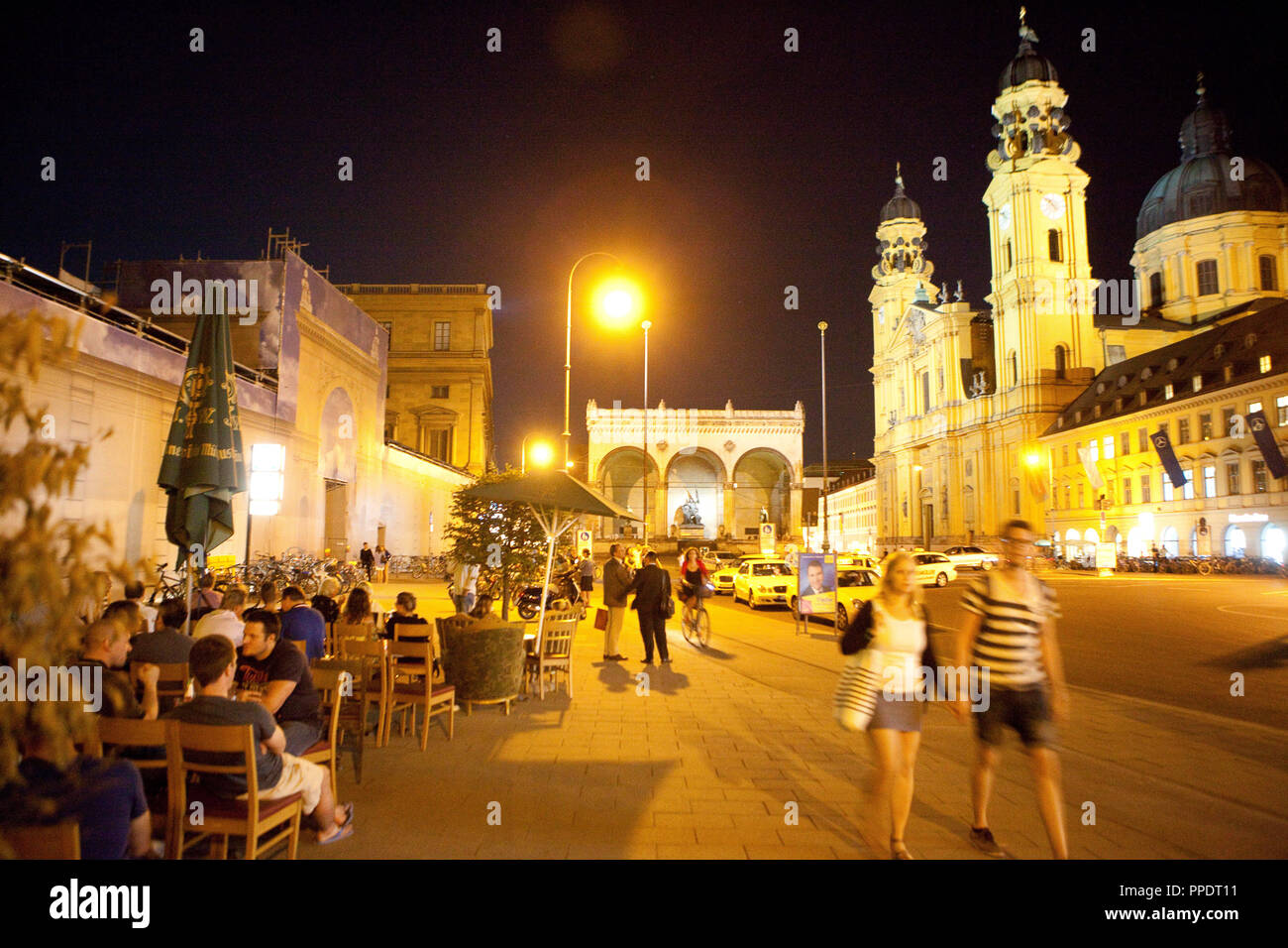 Guests sit on the terrace of Cafe Tambosi on Odeonsplatz on a warm summer evening. In the background, the illuminated Theatinerkirche (Theatine Church) and the Feldherrnhalle. Stock Photo