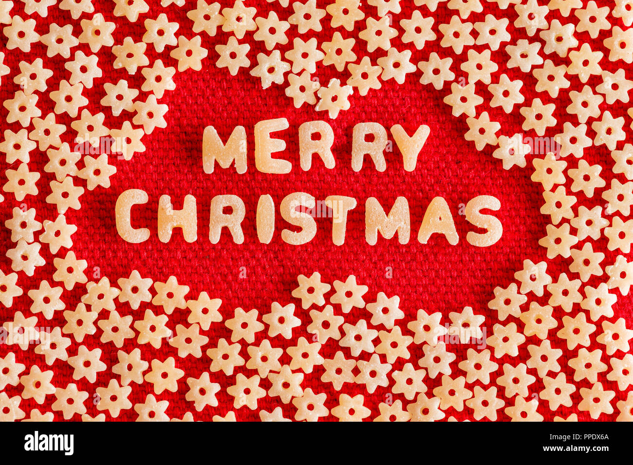 Creative Merry Christmas card design made with dried pasta letters surrounded by a pattern of random scattered stars on a woven red fabric Stock Photo