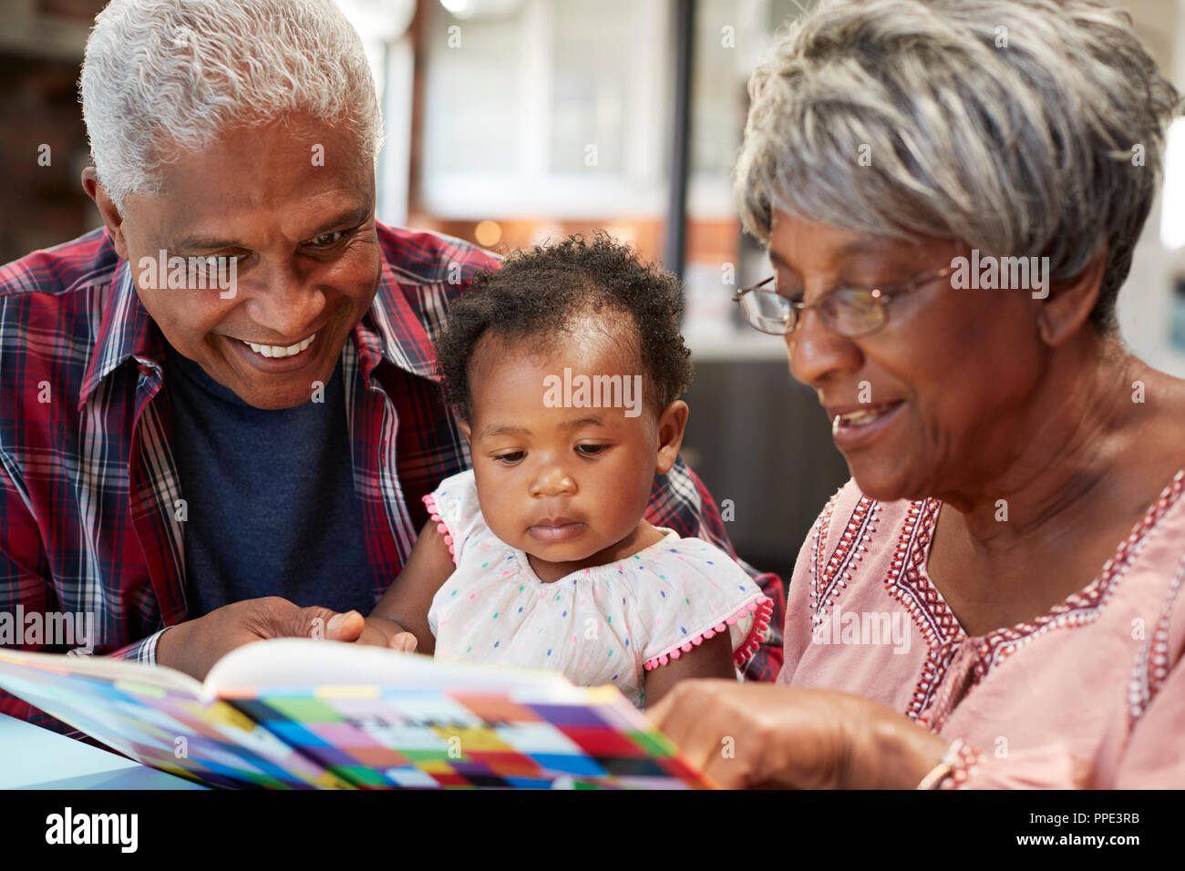 Grandparents Reading Book With Baby Granddaughter At Home Stock Photo
