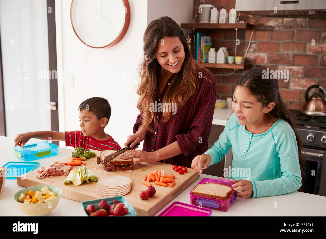 Children Helping Mother To Make School Lunches In Kitchen At Home Stock Photo