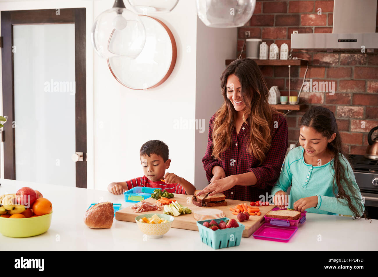 Children Helping Mother To Make School Lunches In Kitchen At Home Stock Photo