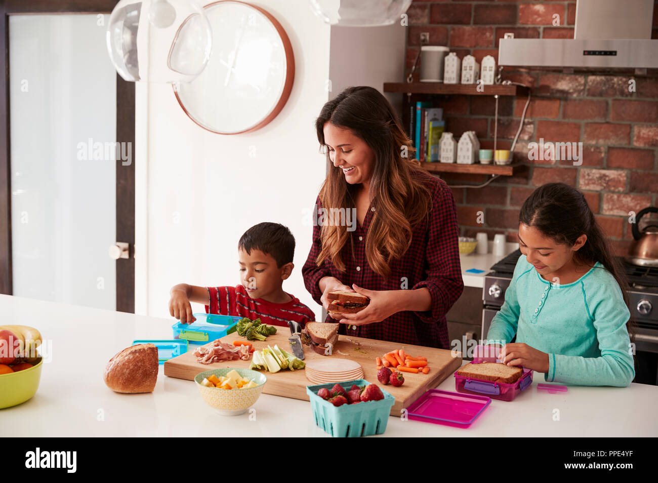 Children Helping Mother To Make School Lunches In Kitchen At Home Stock Photo