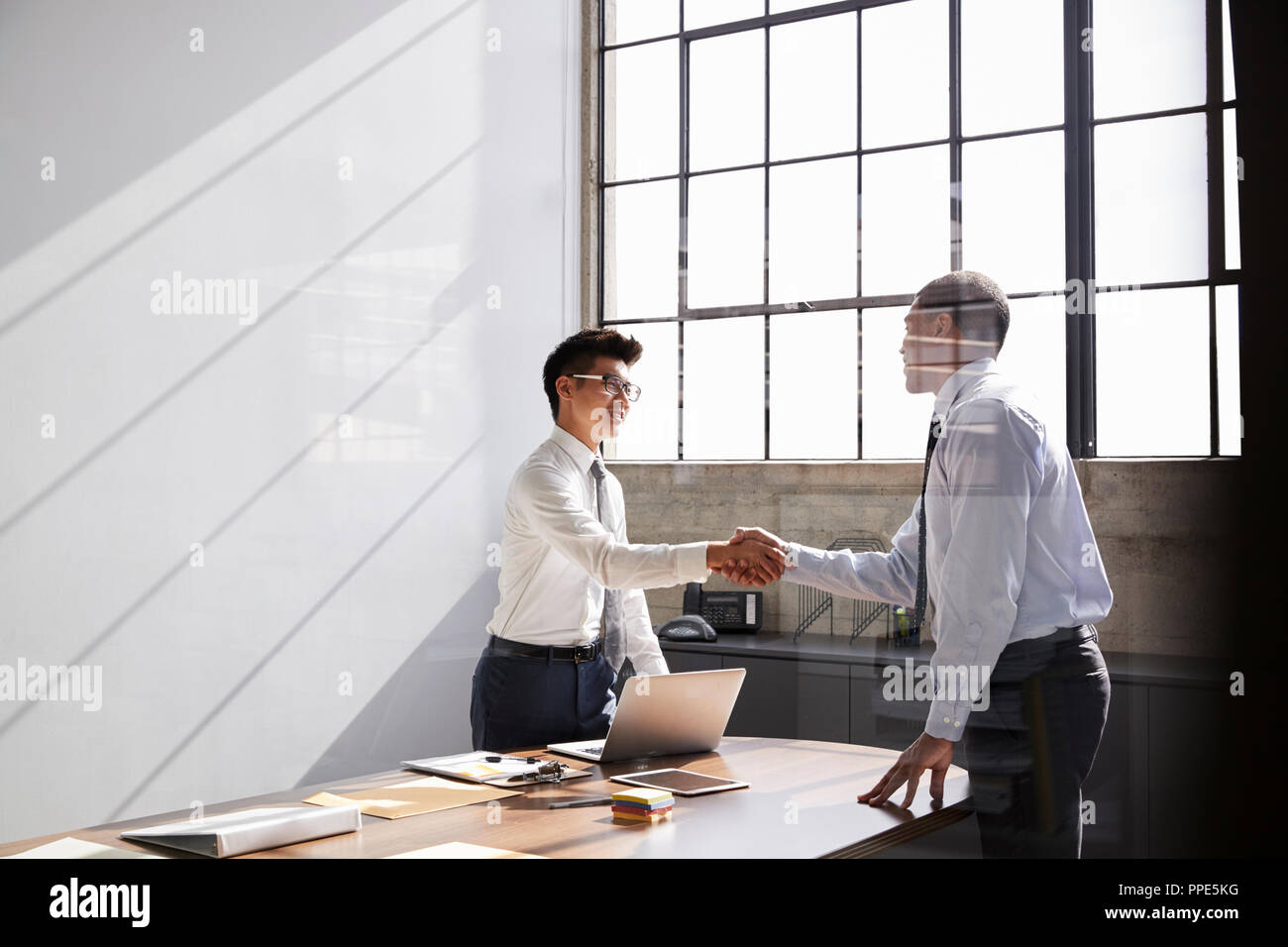 Two businessmen stand shaking hands across desk in an office Stock