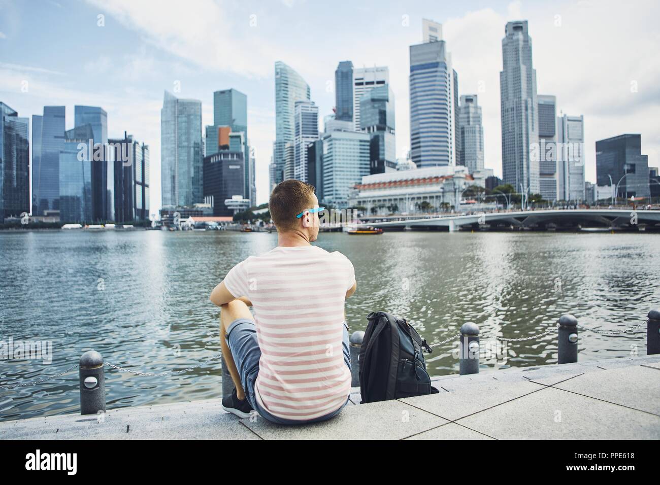 Young man (traveler) with backpack sitting on waterfront against downtown and financial district of Singapore. Stock Photo