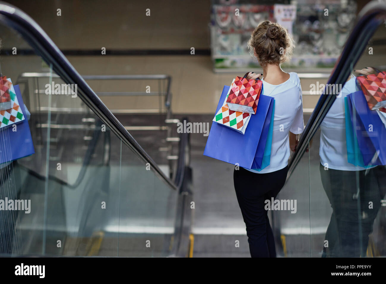 View from back of female customer with shopping bags driving down on escalator in mall. Curly lady enjoying good shopping and new purchases. Concept of buying and pleasure. Stock Photo