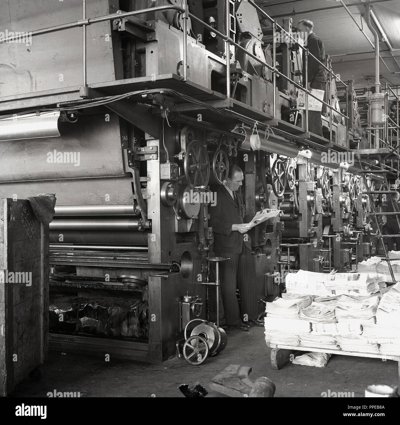 1950s, a suited manager stands underneath the giant printing presses with an operator above, while checking the magazines that have been printed at HWV printers, Aylesbury, Bucks, England, UK. In 1963 the company would become the Britihs Printing Corporation one of the largest printing businesses in Britain, later taken over by Robert Maxwell. Stock Photo