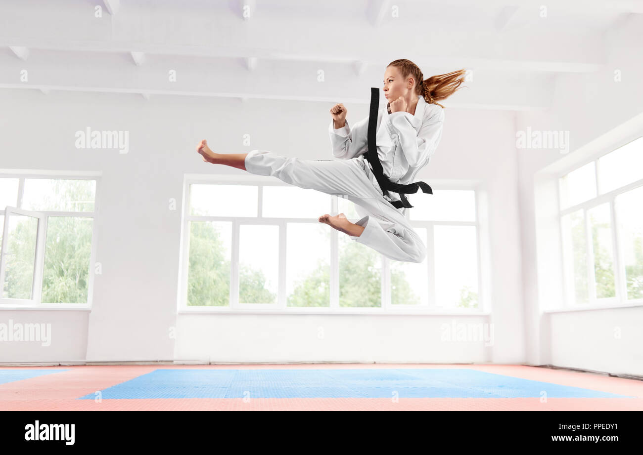 Sporty woman in white kimono with black belt jumping and performing kick. Young girl confidently doing complex technique elements karate with serious facial expression. Healthy lifestyle concept. Stock Photo