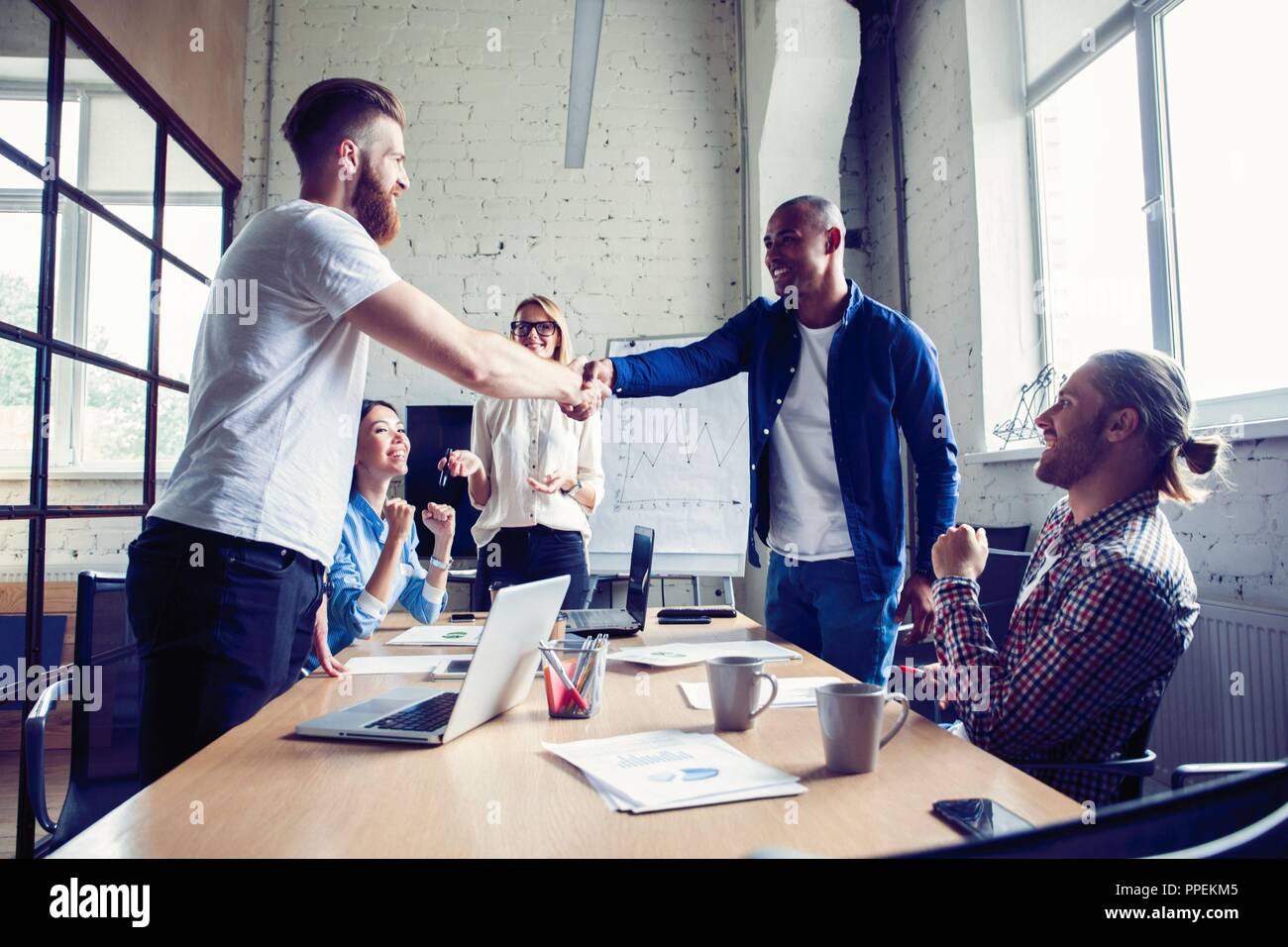 New business partners. Young modern colleagues in smart casual wear shaking hands and smiling while sitting in the creative office. Stock Photo