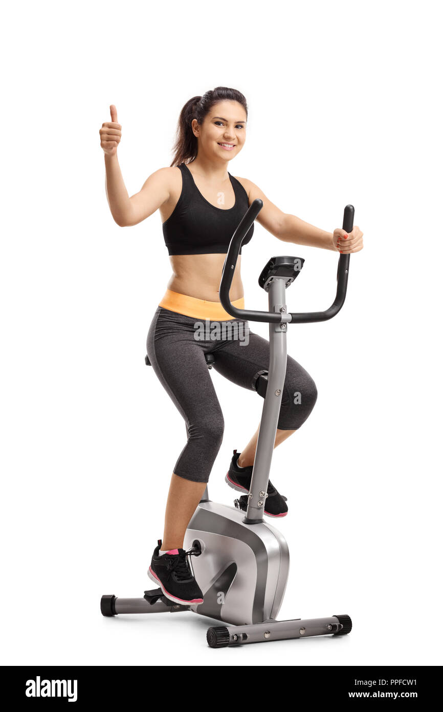 Full length portrait of a young female riding a stationary bike and showing thumb up isolated on white background Stock Photo