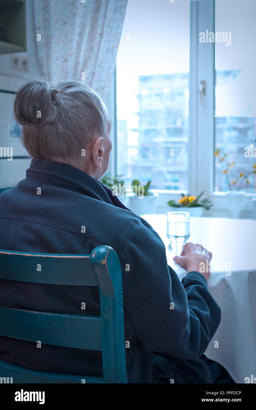 Old woman sitting alone at her kitchen table looking out of the window, blue filter effect, concept of old-age depression, loneliness Stock Photo