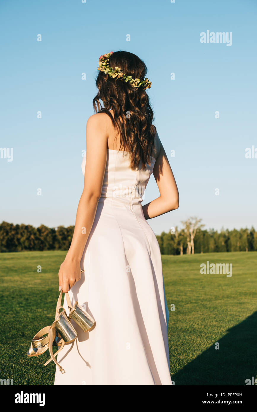 back view of beautiful tender young bride holding shoes on green lawn in park Stock Photo