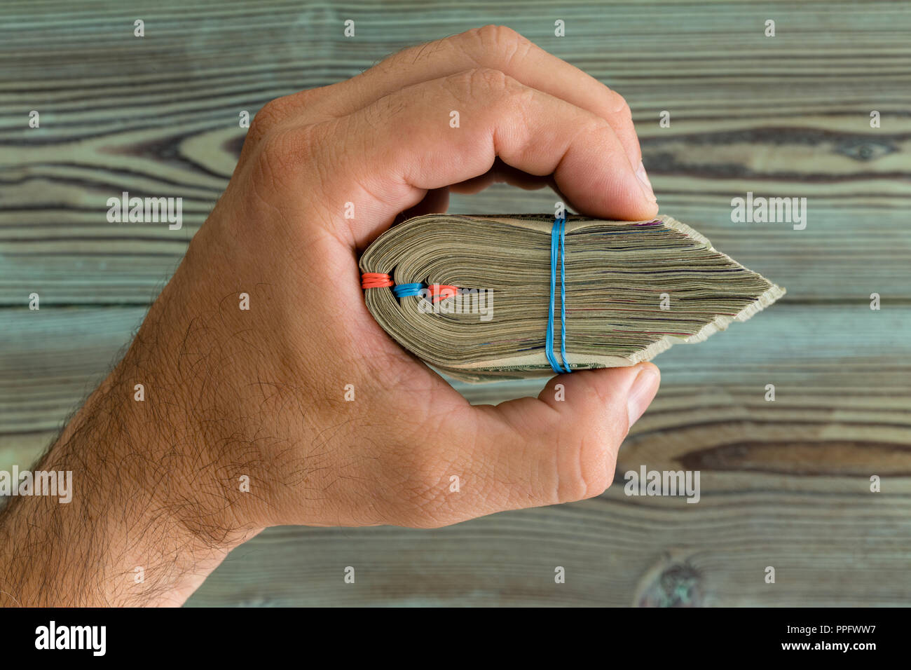 Man holding a thick wad of banknotes tied with a rubber band in his hand over wood in a