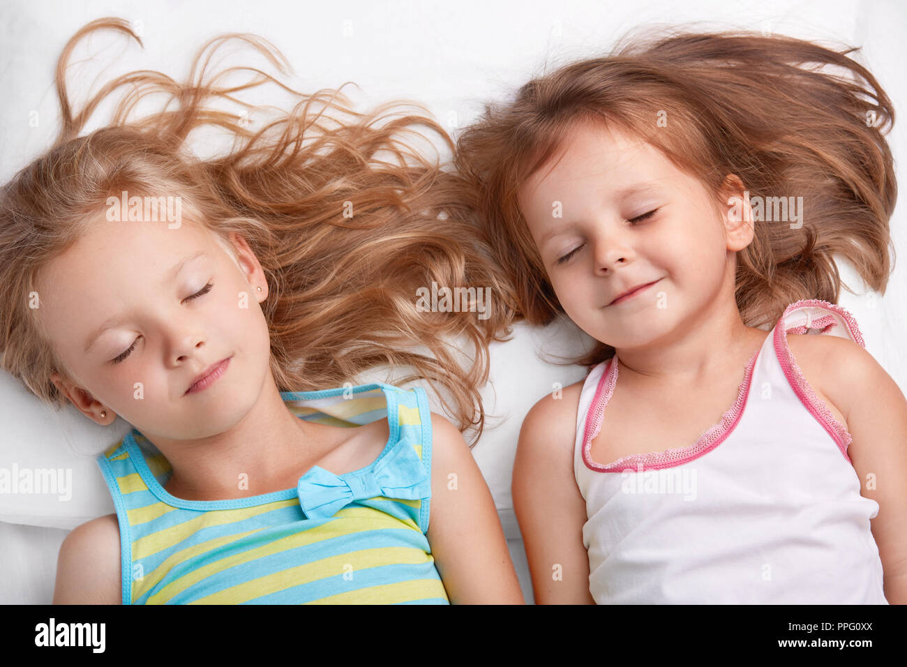 Top view of pleased attractive small girls or sisters smile together, sleep in bed, lie closely to each other, see pleasant dreams, dressed in nightwe Stock Photo
