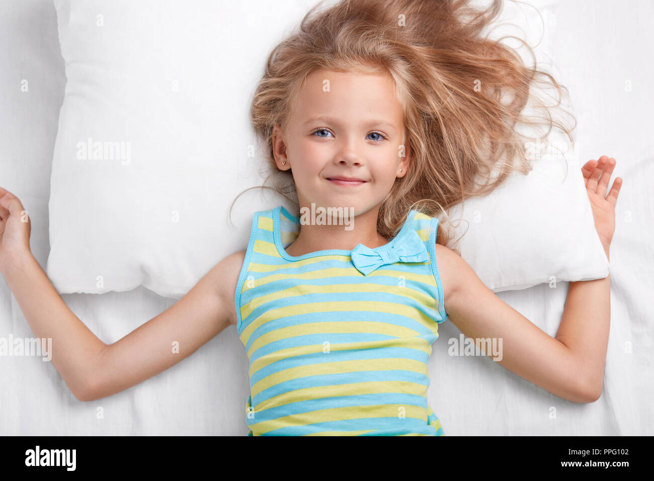 Children, bedding and awakening concept. Pleased small European girl with charming tender smile, long light hair, dressed in pyjamass, lies on white p Stock Photo