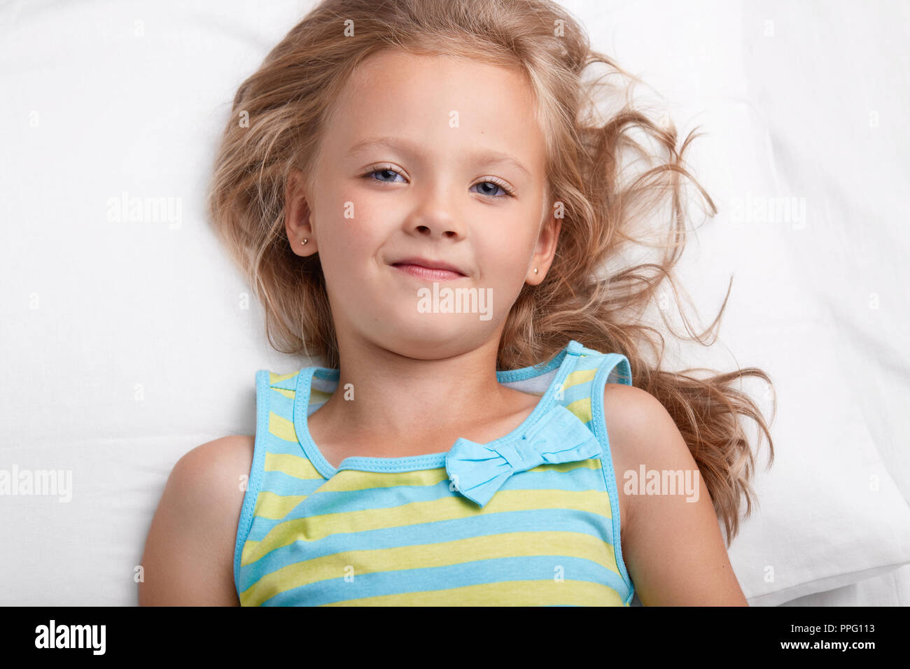 Close up shot of charming small kid with healthy skin, light hair, blue appealing eyes, healthy soft skinm wears pyjamass, lies on white bedclothes in Stock Photo