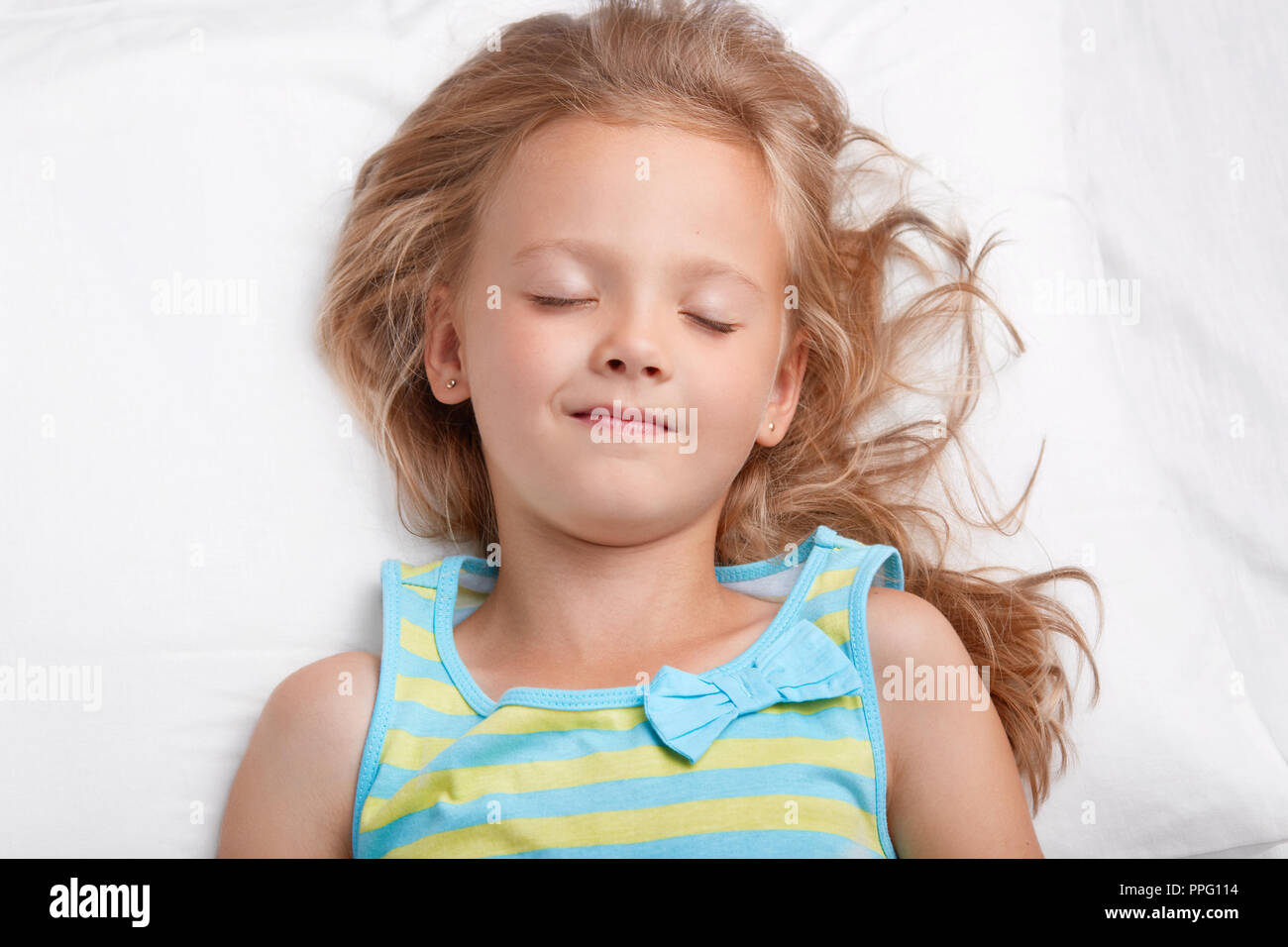 Healthy appealing little girl with long messy hair, keeps eyes shut, dressed in pyjamass, has