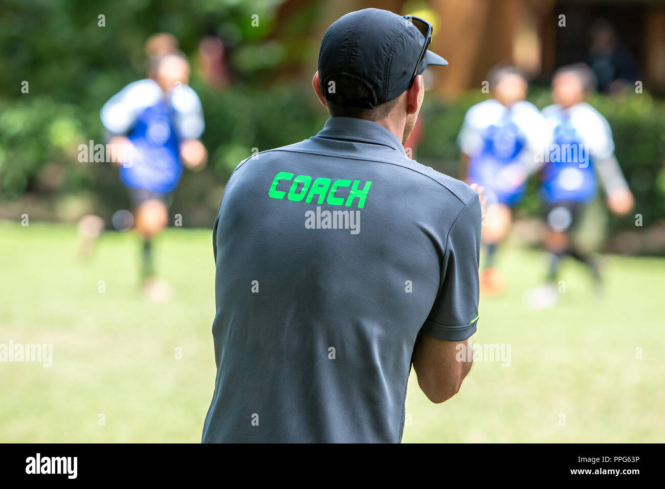 Male soccer or football coach in gray shirt with word COACH written on back, standing on the sideline watching his team play, good for sport or coachi Stock Photo