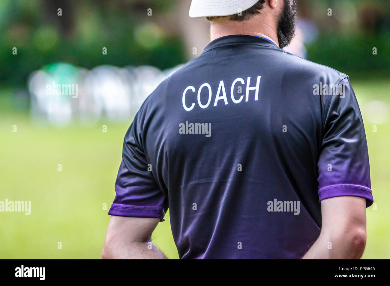 Back view of male soccer or football coach in dark shirt with word COACH written on back, standing on the sideline watching his team play, good for sp Stock Photo