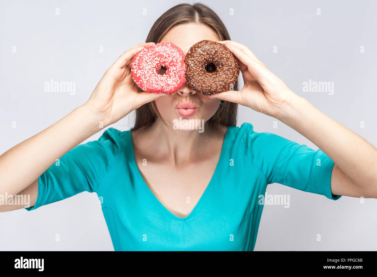 Positive young beautiful girl in blue blouse standing, holding, covering eyes with pink and chocolate donuts,looking through donuts like making glasse Stock Photo