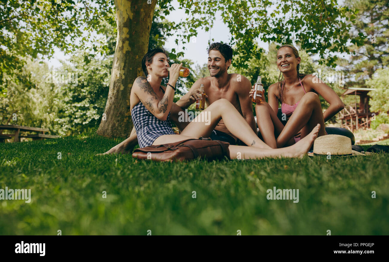 Two women and a man sitting in park together holding beer. Friends having fun drinking beer sitting in park. Stock Photo