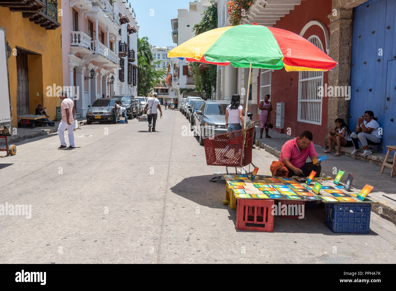 Cartagena, Columbia--April 21, 2018. People go about their business on a side street in Cartagena while an artist paints  under a colorful umbrella. Stock Photo