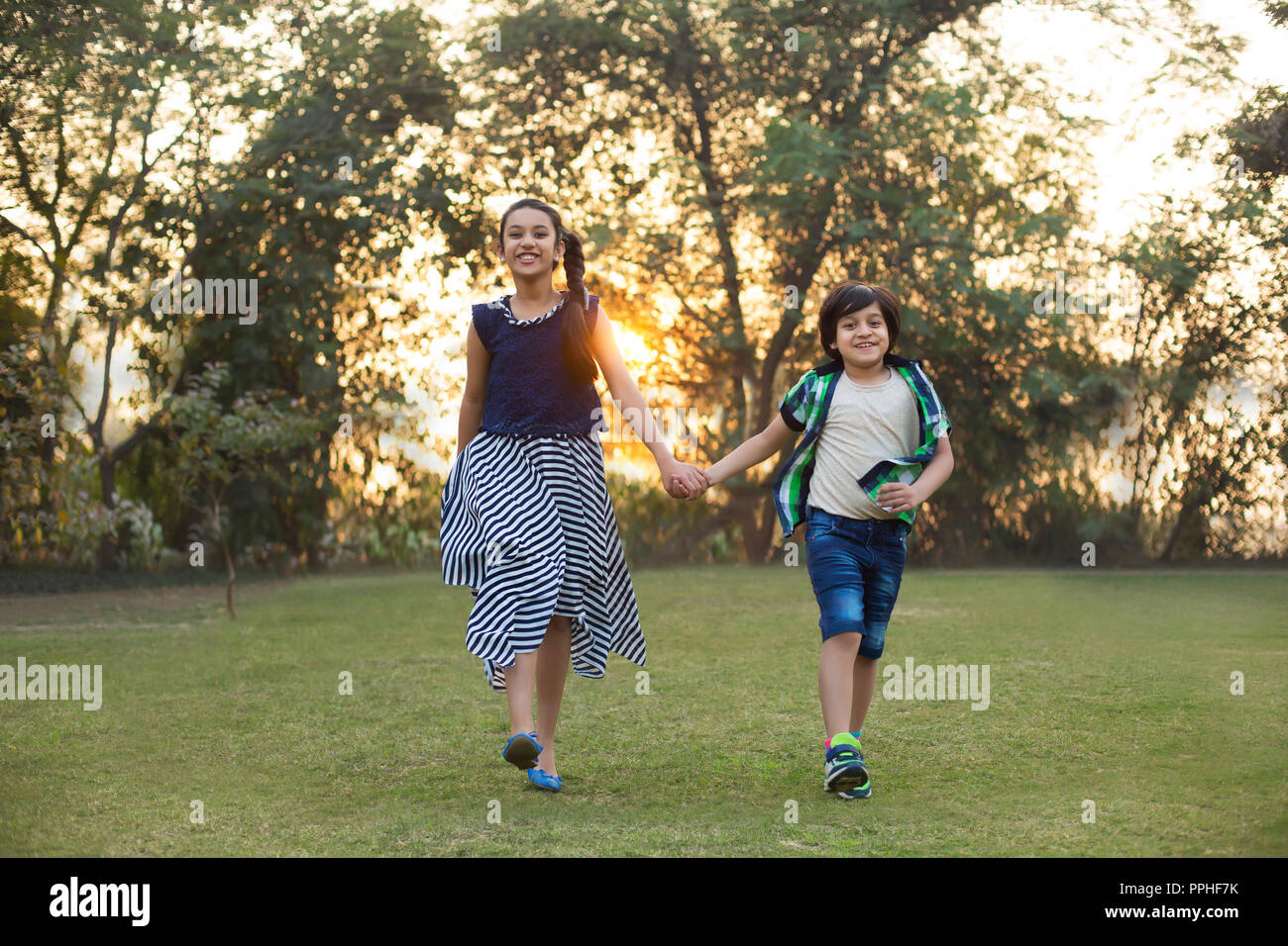 Happy brother and sister walking in park holding hands with sunlight in the background. Stock Photo
