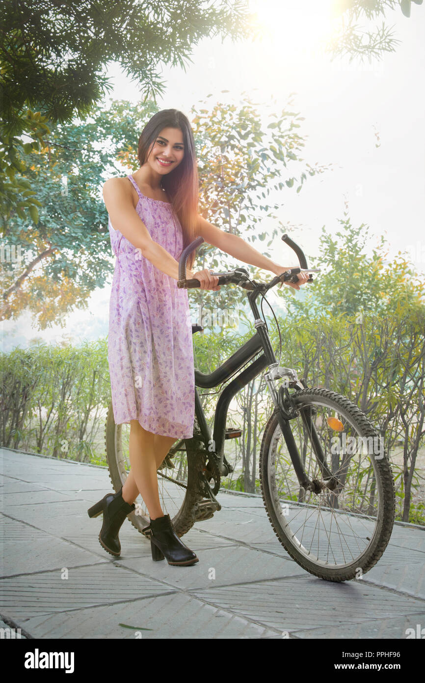 Beautiful smiling young woman walking on the pavement in a park holding a bicycle. Stock Photo