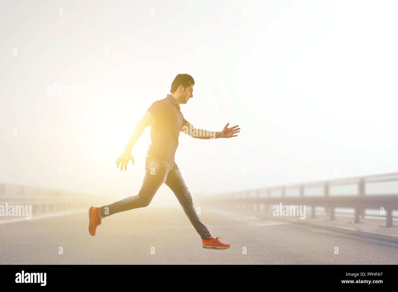 Young man leaping joyfully in mid air across the road with sun in the background. Stock Photo