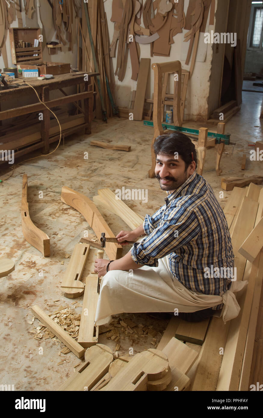 Smiling carpenter working on wood with a chisel and hammer sitting in his workshop. Stock Photo