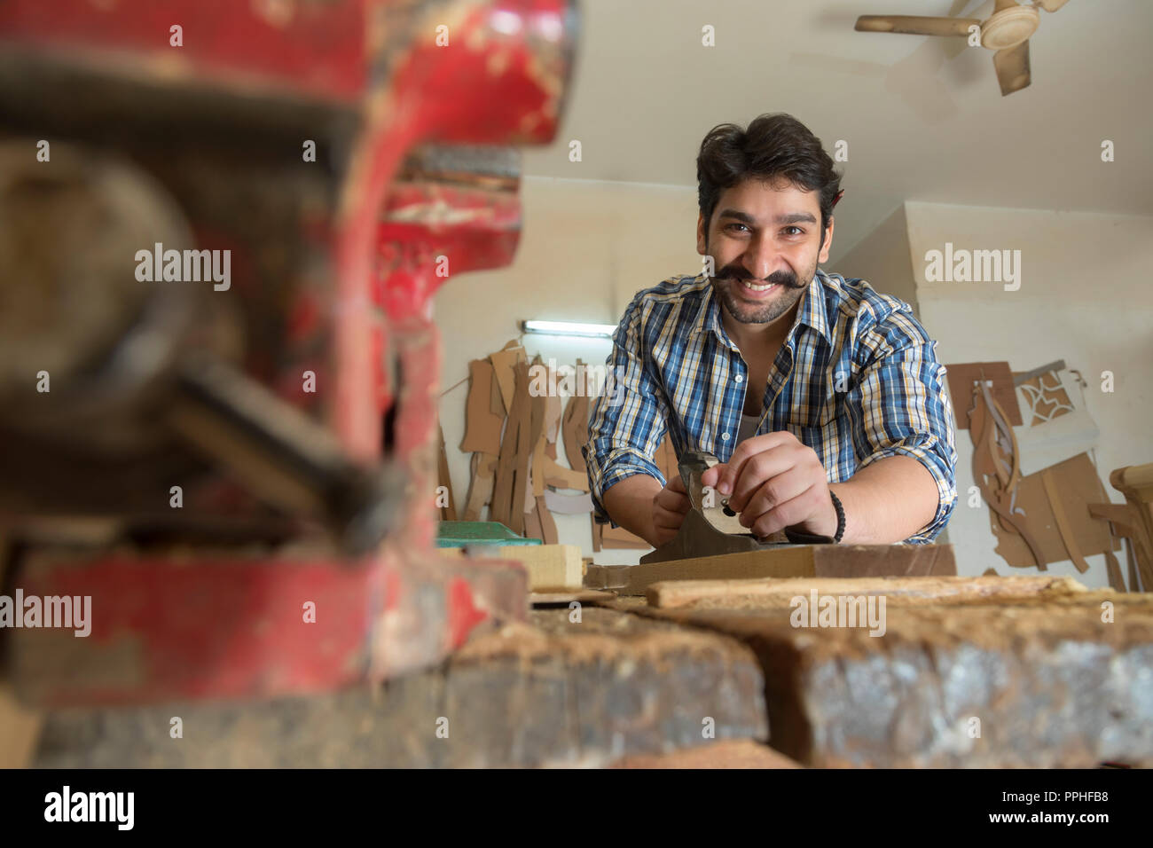Smiling carpenter using a scraper blade to smoothen wooden log with a metal clamp in the foreground. Stock Photo
