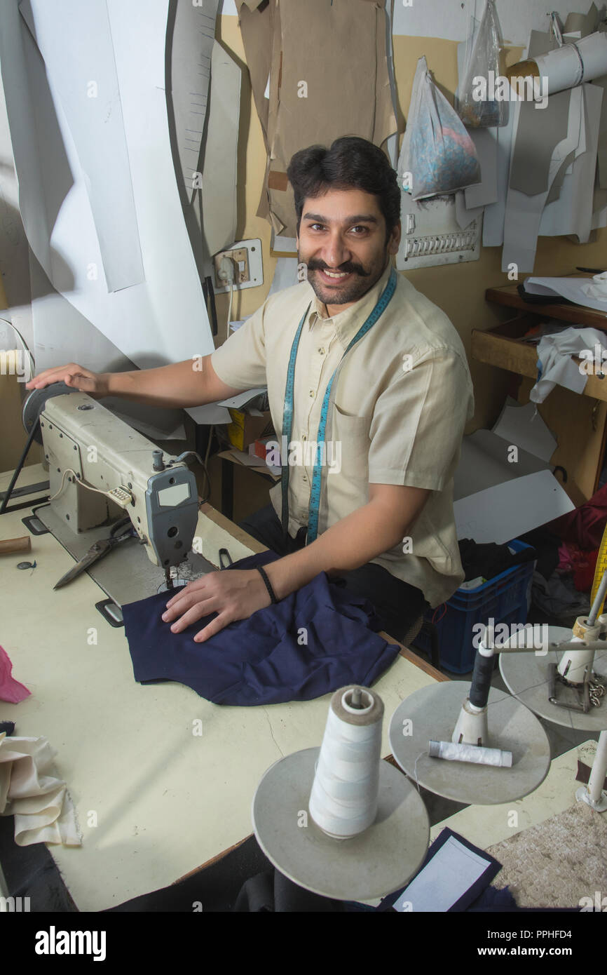 Happy looking tailor working on sewing machine stitching a cloth in his small workshop. Stock Photo