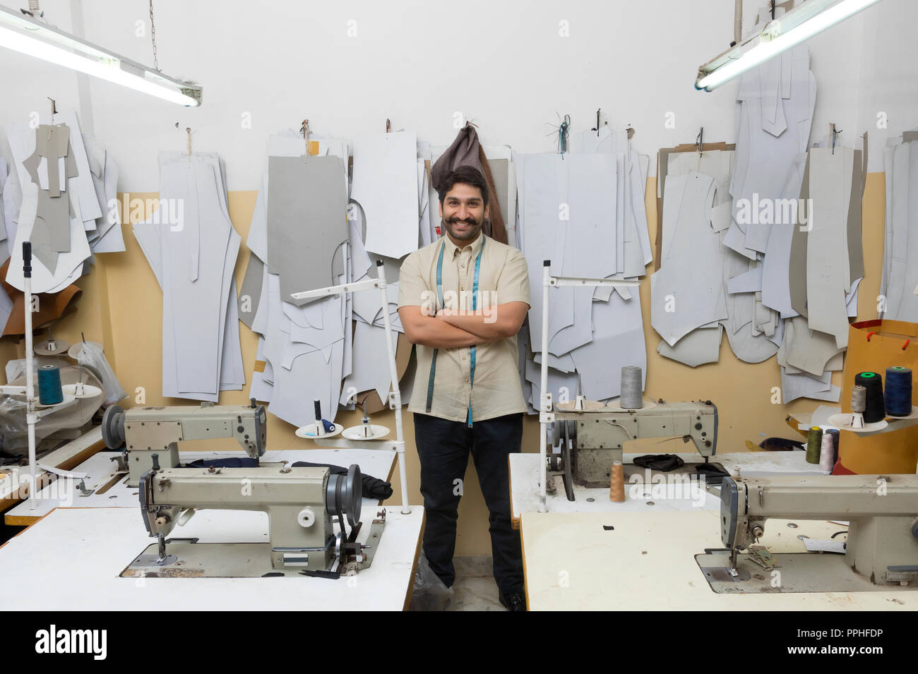 Smiling tailor standing with arms crossed in his workshop with sewing machine around and sample cuttings hanging in the background. Stock Photo