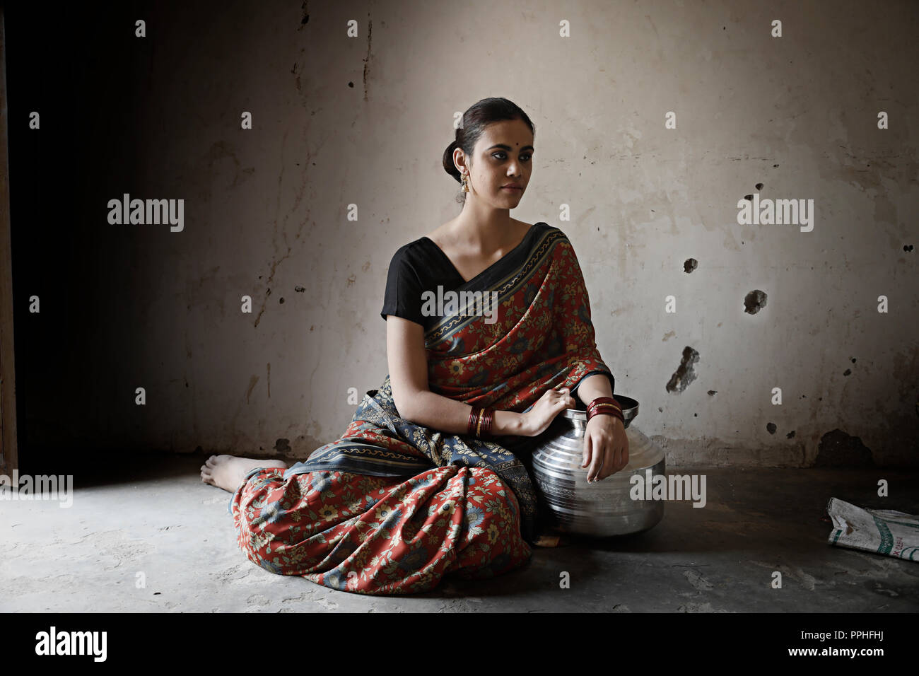 Beautiful village woman sitting on the floor with a steel pot in an old room looking away. Stock Photo