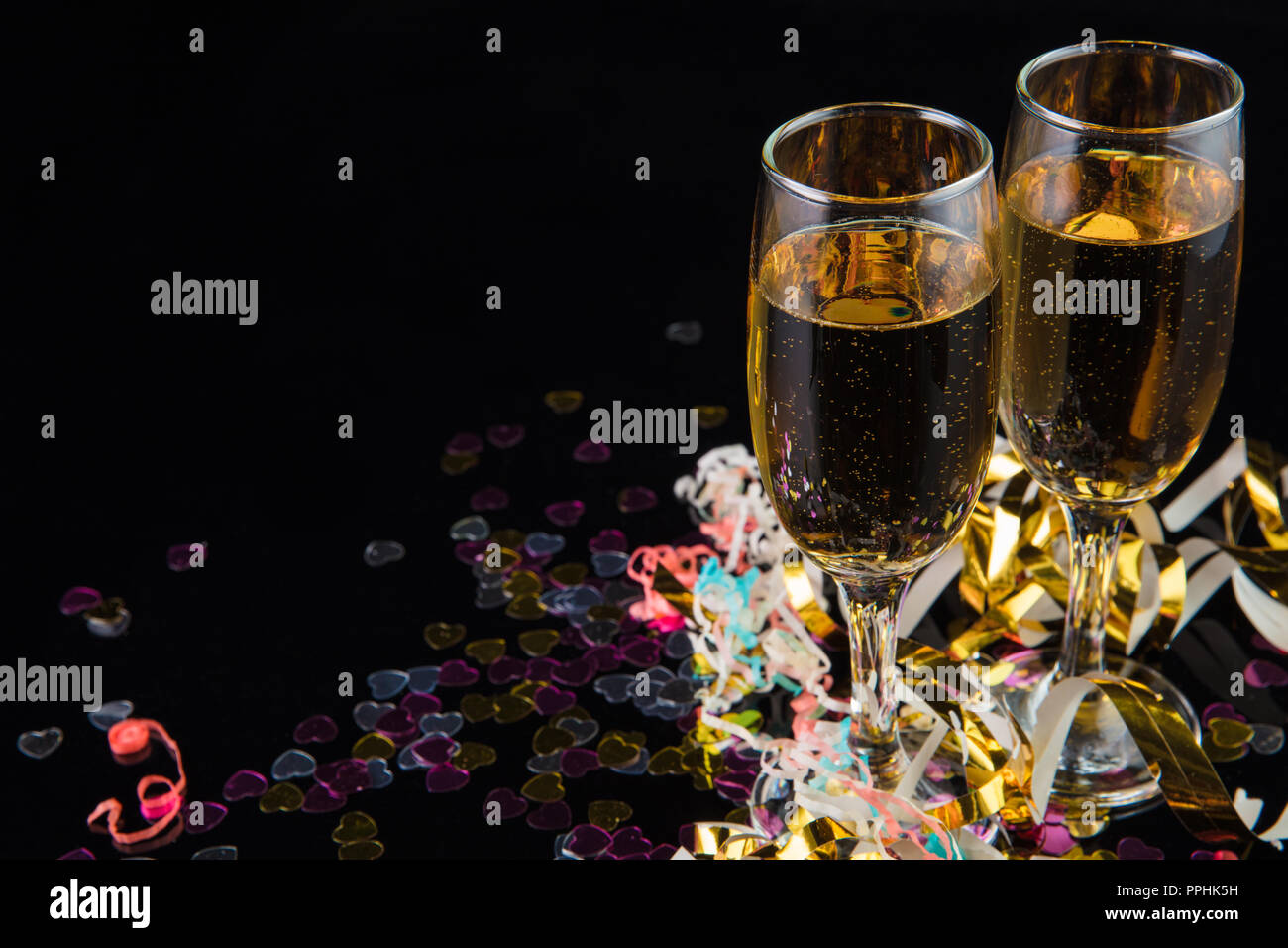 two glasses of champagne on a shiny black surface with confetti against a black background. Stock Photo