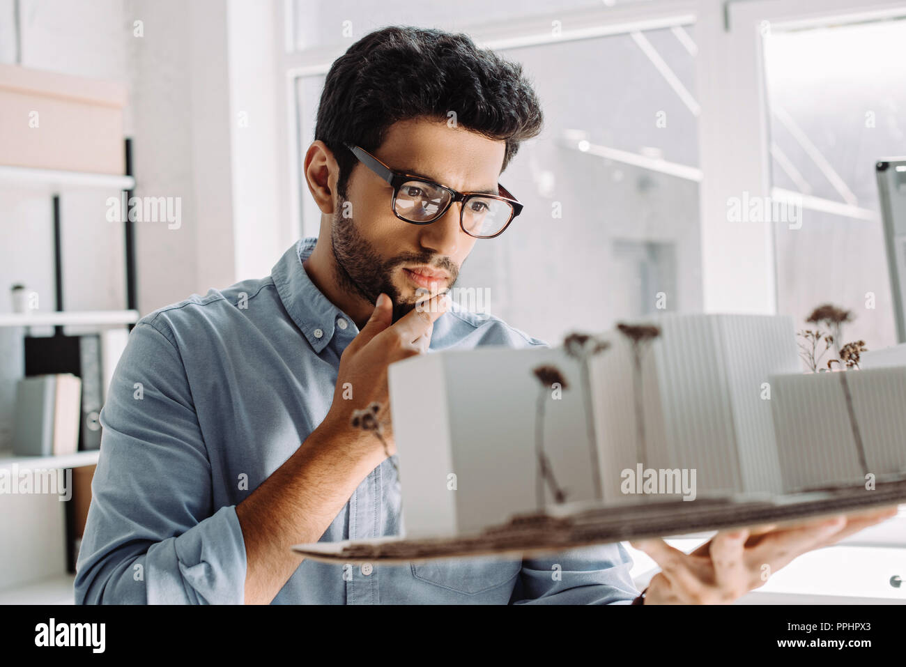 pensive architect in glasses touching chin and looking at architecture model in office Stock Photo