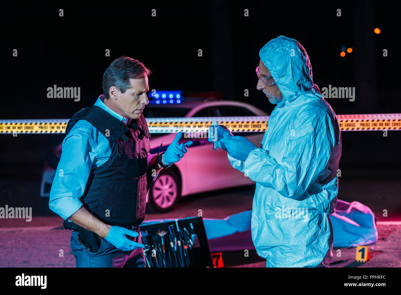 male criminologist in protecitve suit showing evidence to mature policeman at crime scene with corpse in body bag Stock Photo
