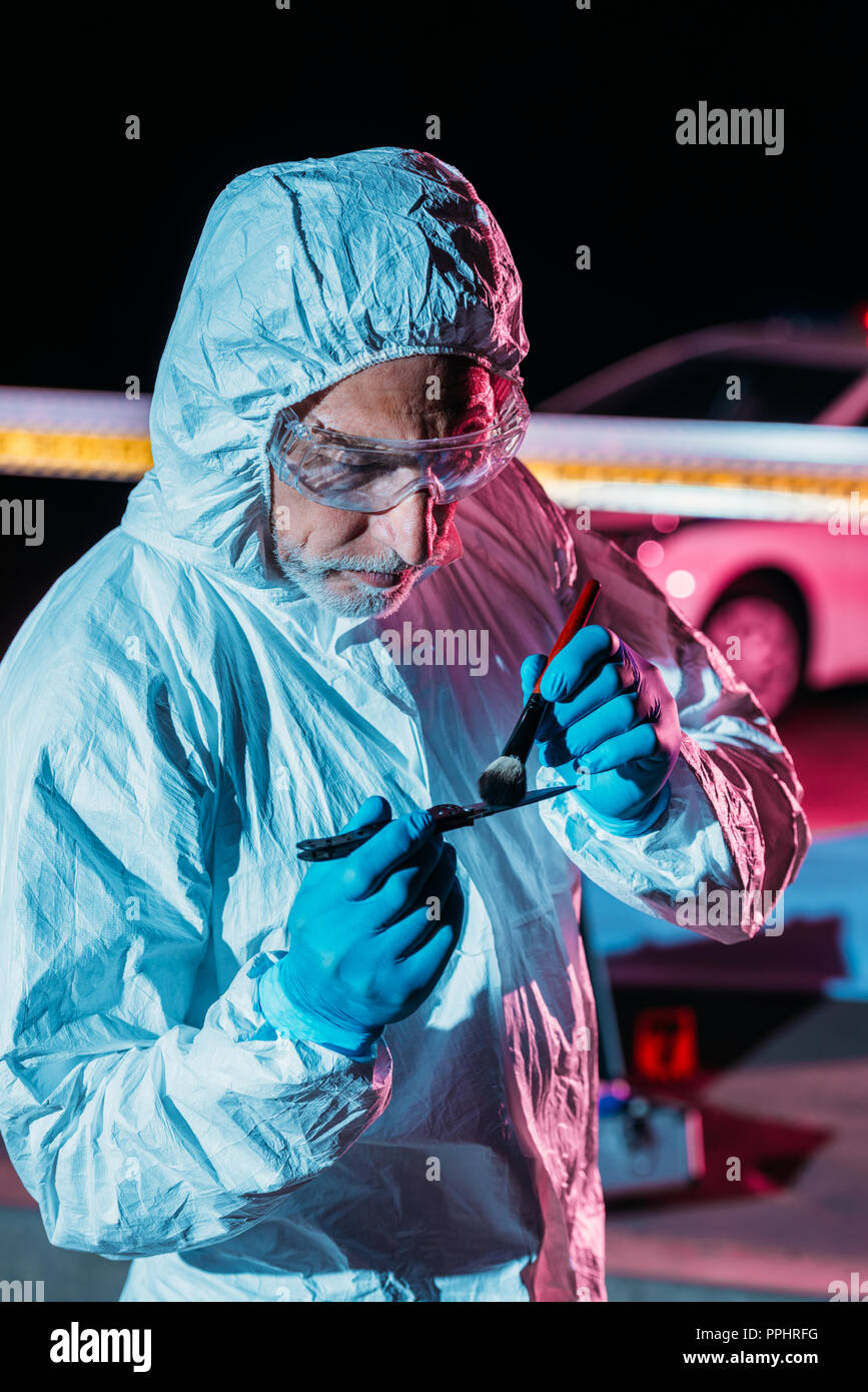 male criminologist in protective suit and latex gloves taking fingerprints from knife at crime