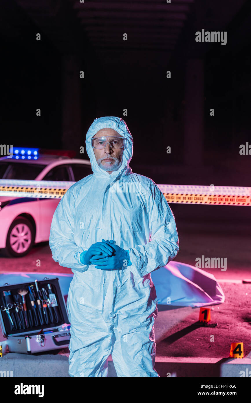 male criminologist in protective suit and mask looking at camera near crime scene with corpse in body bag and case with investigation tools Stock Photo