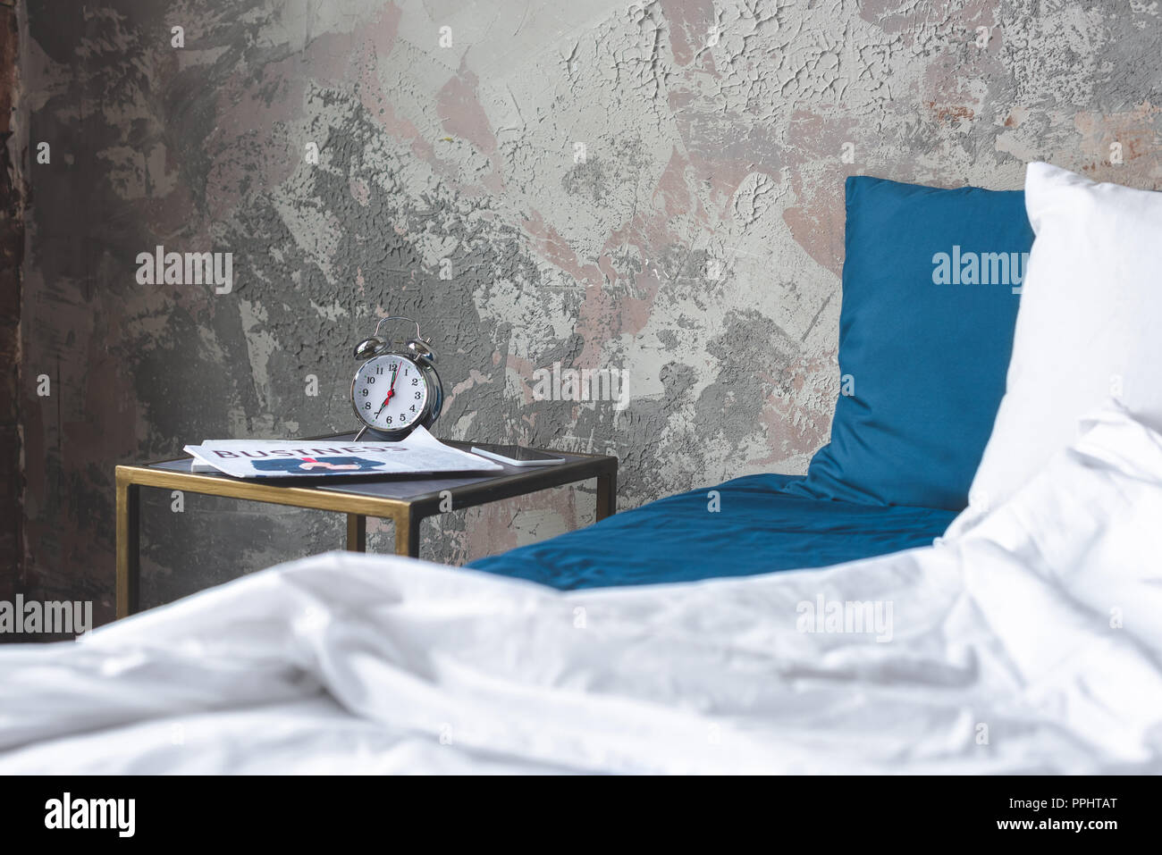 messy bed in loft bedroom with vintage alarm clock and newspaper on bedside table Stock Photo