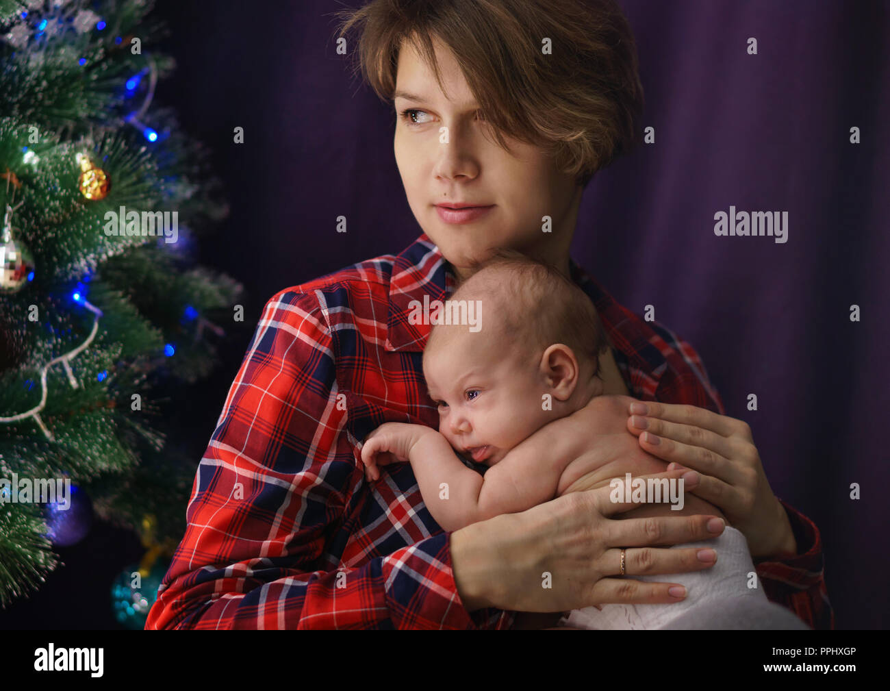 Family celebration. Portrait of young mother and newborn baby. In background Christmas tree. Happiness of motherhood. Stock Photo
