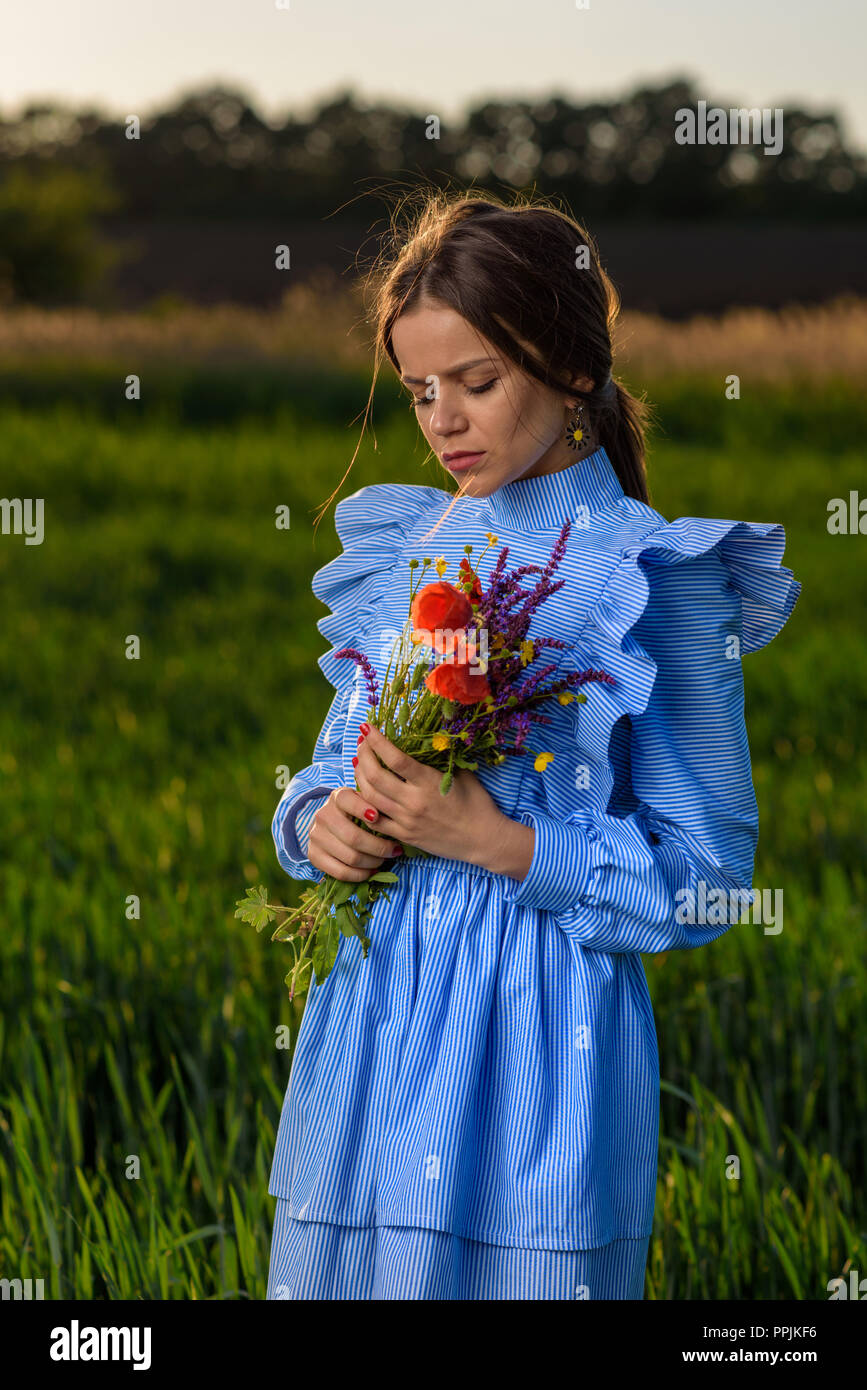 Young woman in blue and white striped dress is holding with two hands a bouquet of summer flowers with her eyes closed and her head is tilted while st Stock Photo