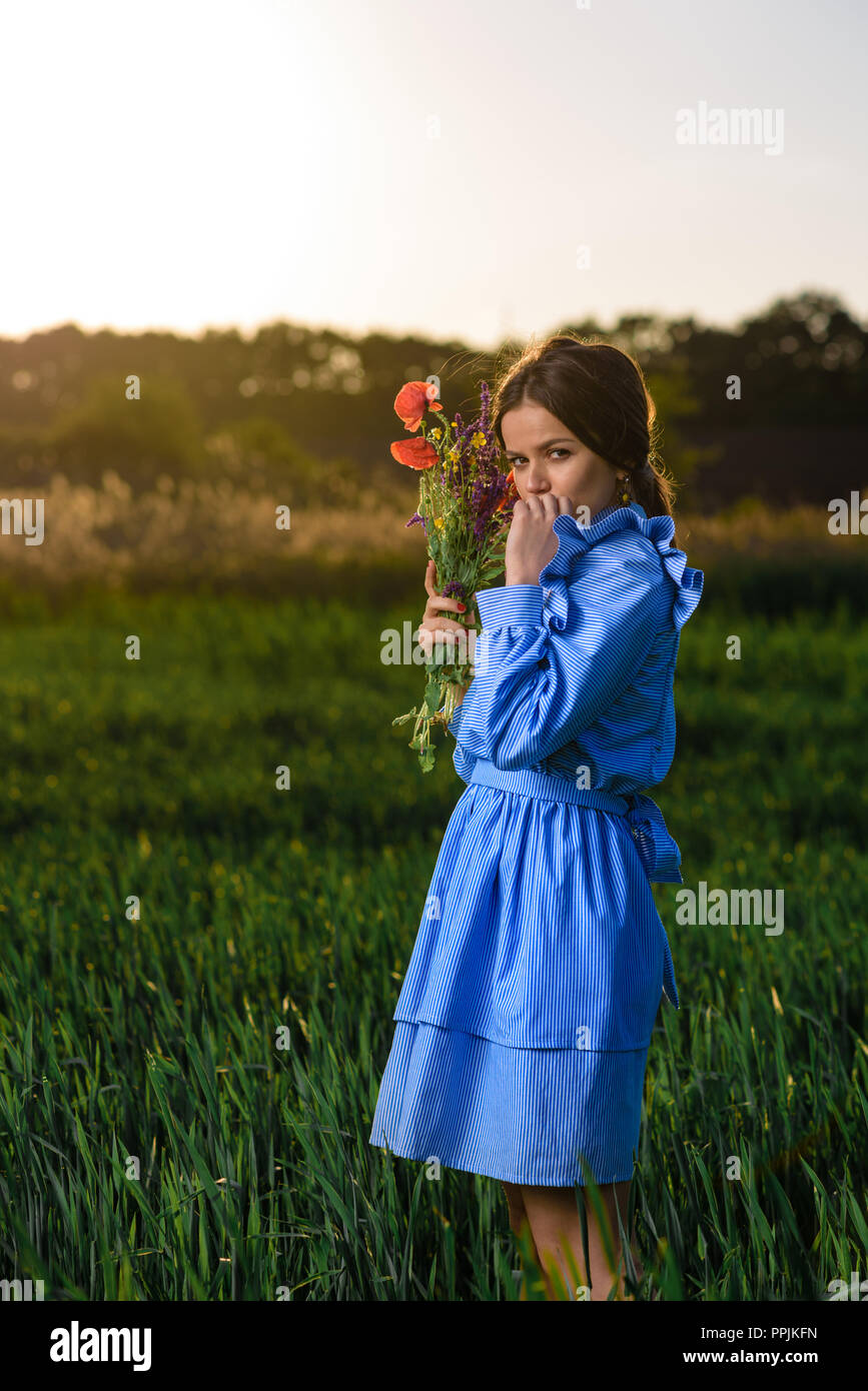 Young woman in blue and white striped dress is holding a bouquet of summer flowers and looking to the camera while standing in the field of green whea Stock Photo