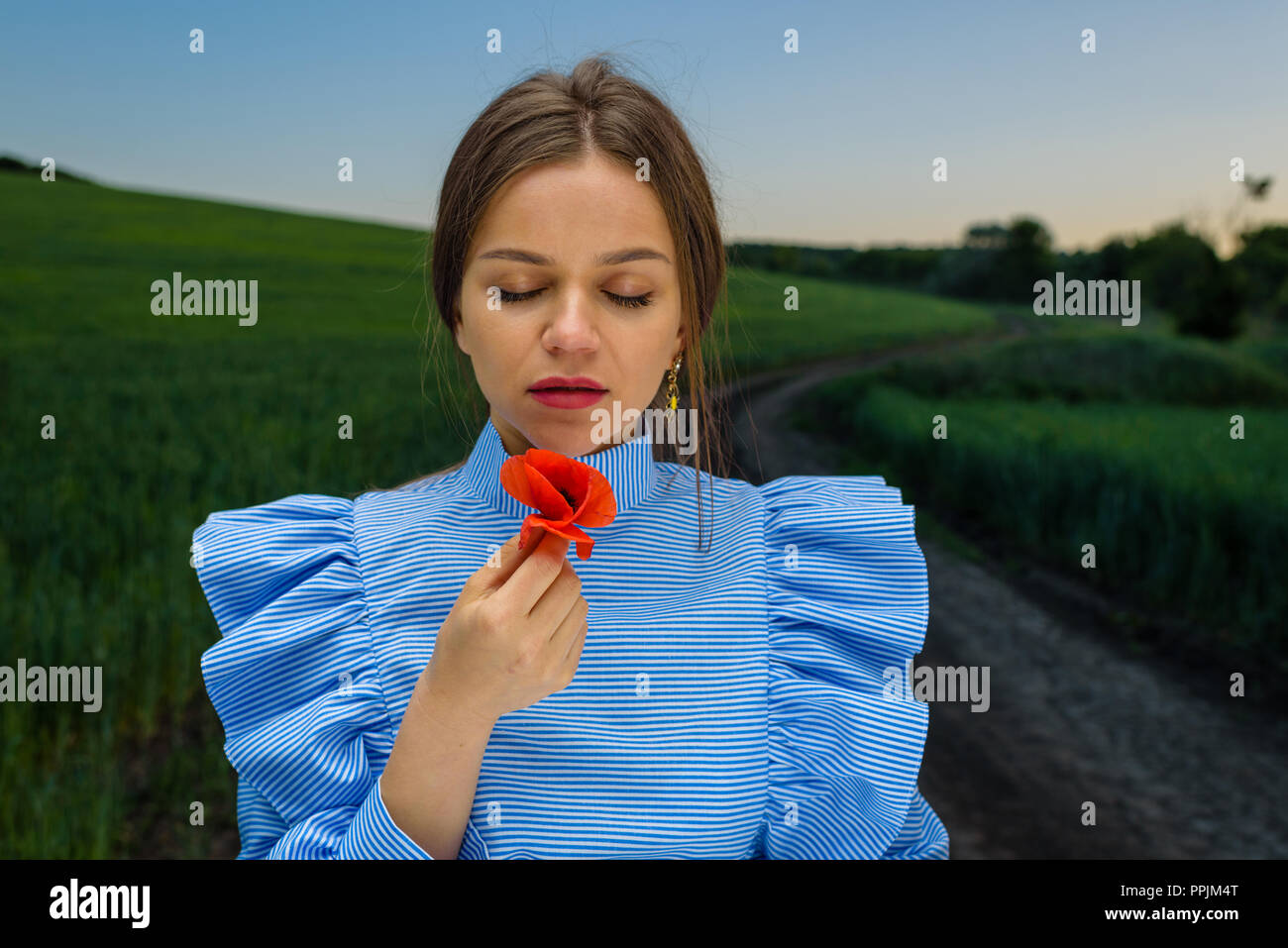Young woman in blue and white striped dress is holding a red poppy flower in her hand with her eyes closed while standing on the country road near fie Stock Photo