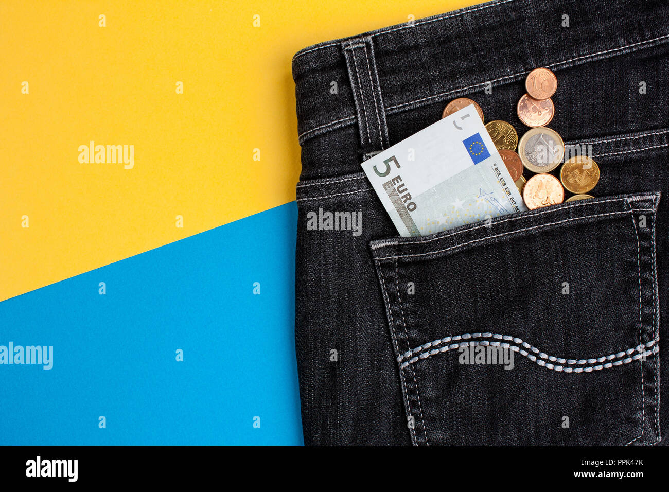 Euro banknote and coins in black jeans back pocket on yellow and blue background Stock Photo