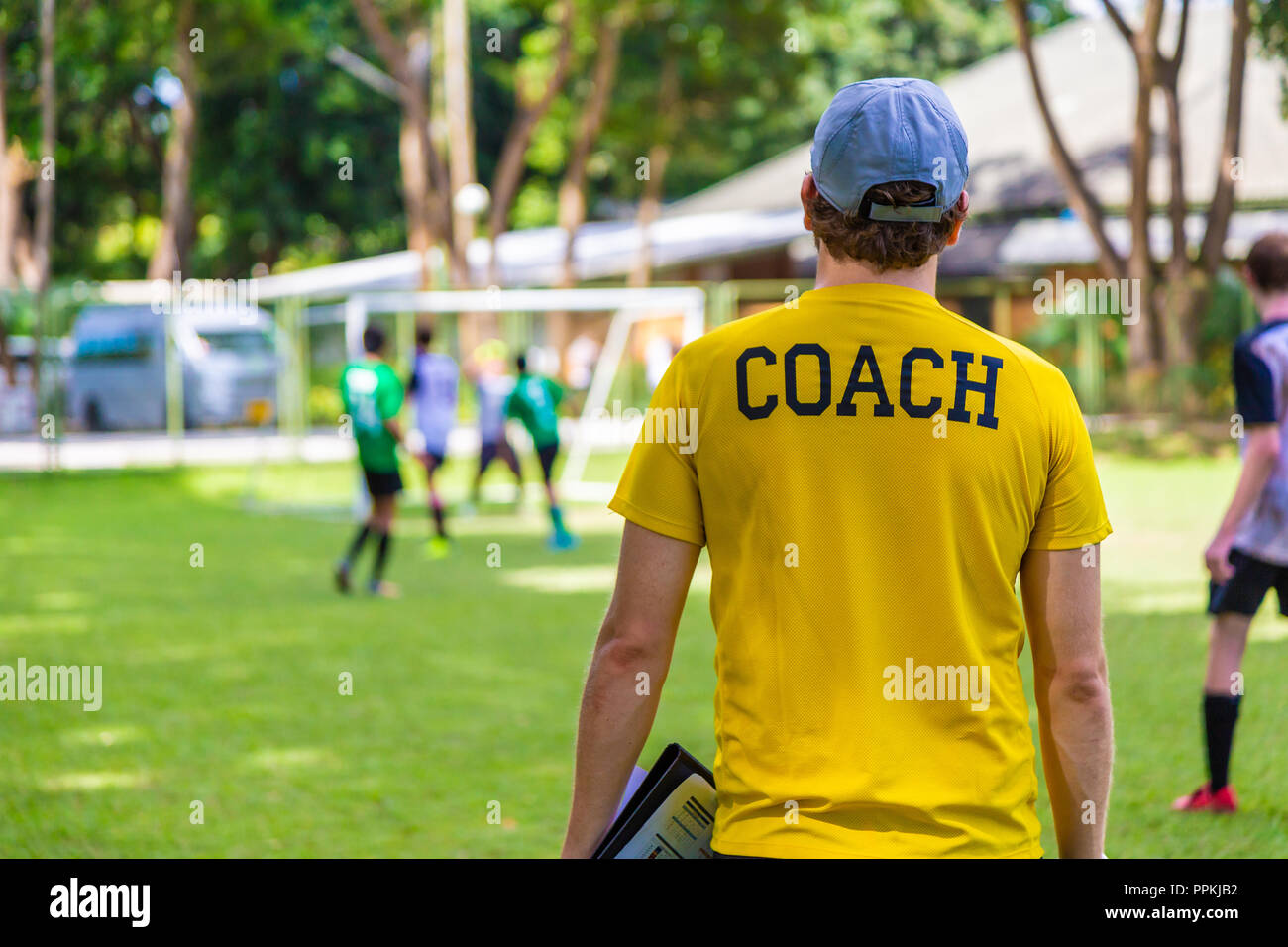 Back view of male soccer or football coach in yellow shirt with word COACH written on back, standing on the sideline watching his team play, good for  Stock Photo