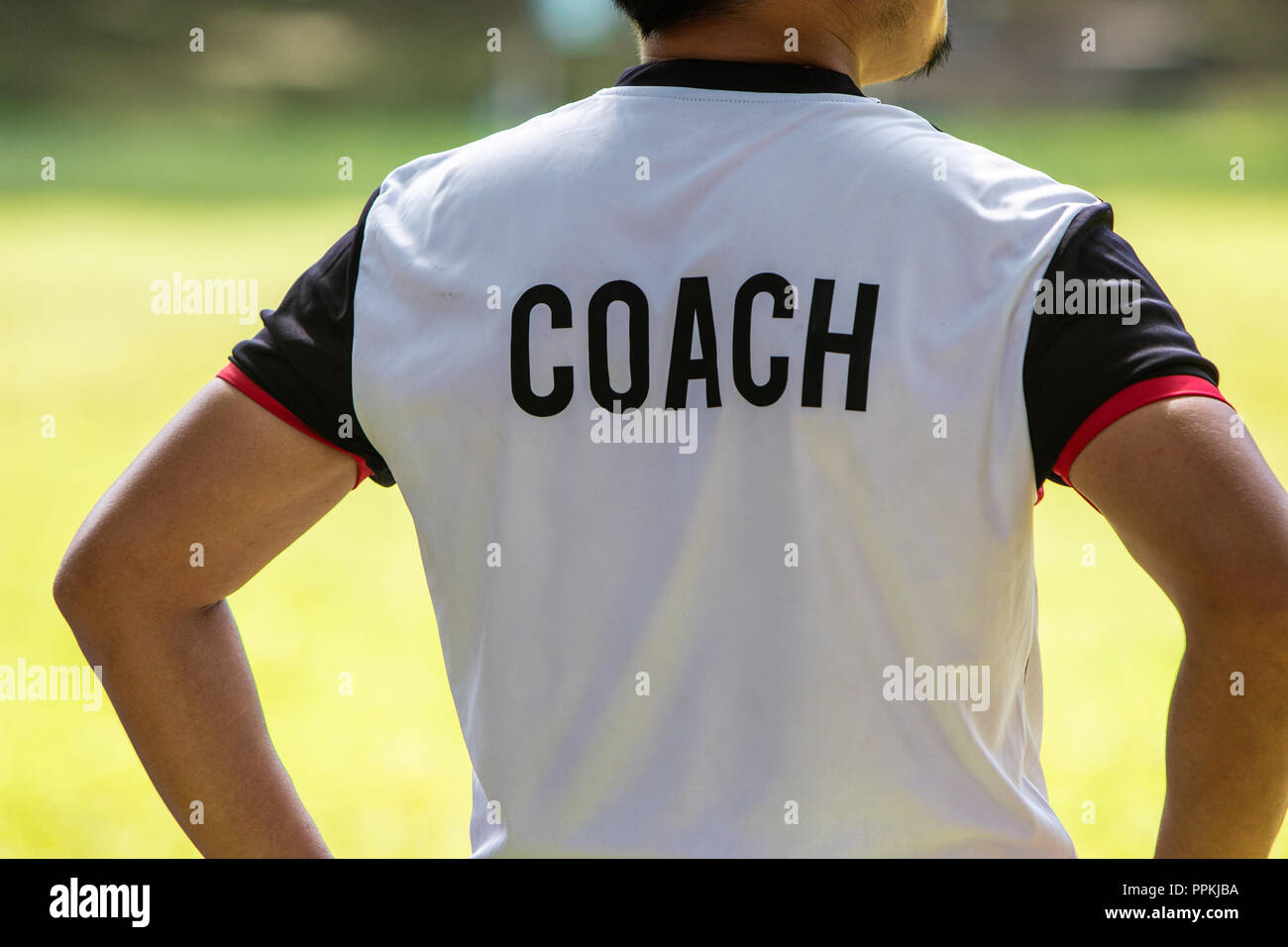 Back view of male soccer or football coach in white shirt with word COACH written on back, standing on the sideline watching his team play, good for s Stock Photo